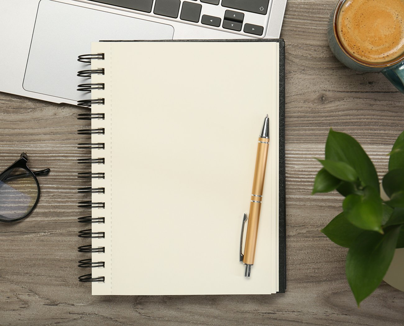 Photo of a wooden desktop on which is places a spiral notebook and pen, glasses, a green mug of coffee and a small, leafy green plant. A laptop keyboard is visible at the top of the image.