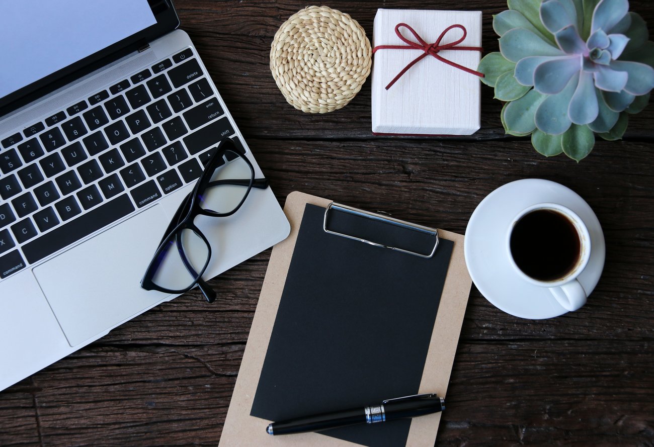 Photo of a creative editorial workspace with laptop, glasses, clipboard and pen, a small raffia pot with a lid, a pile of blank white cards secured in a bow with a red lace, a succulent plant and a black coffee in a white cup