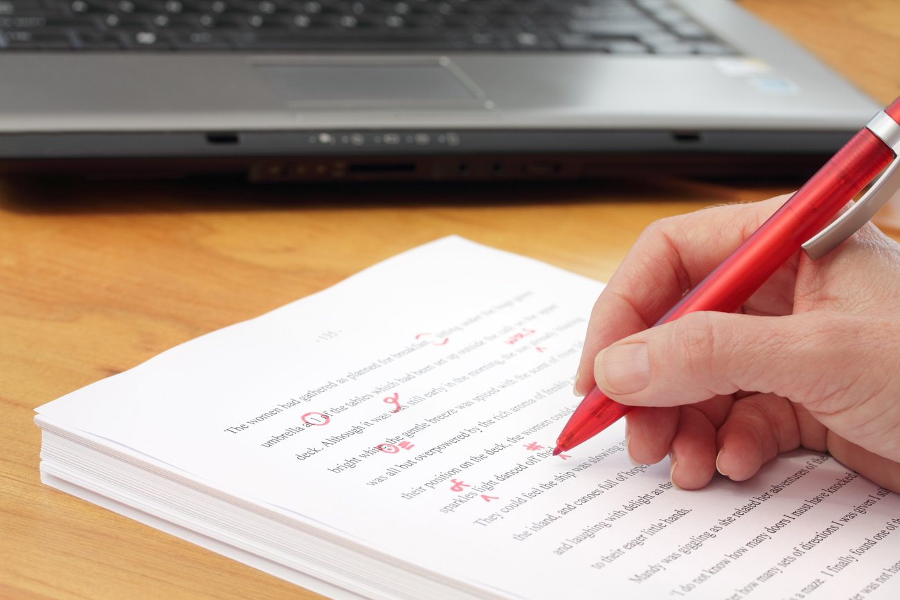 Close-up of a proofreader marking up corrections with a red pen on a printed manuscript. The photo shows the top of a light brown desk with the proofreader’s hand holding the pen over a large pile of printed pages. There’s th